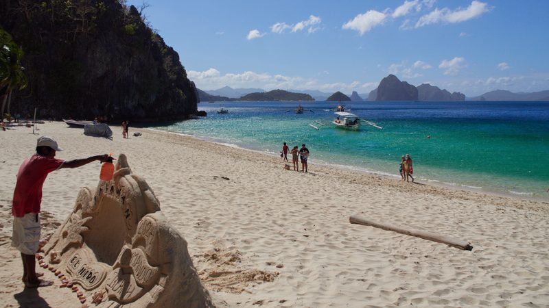 A sand sculptor works on a carved sand sculpture, with outrigger boats moored in turquoise water and limestone